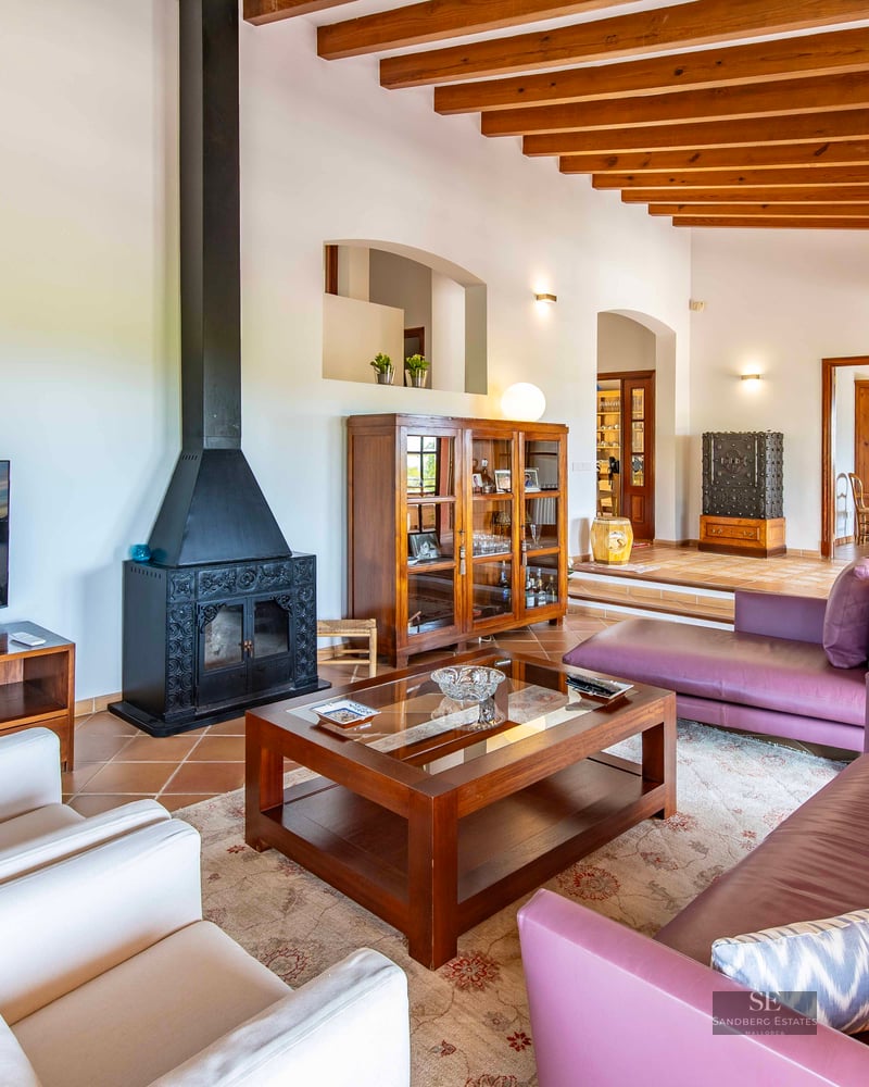 Living room featuring purple and white sofas, a central coffee table, a black fireplace, and exposed wooden ceiling beams.