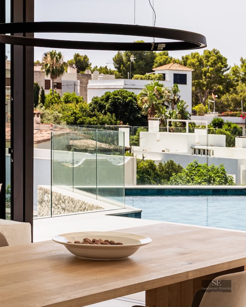 A minimalist dining room with a wooden table and glass walls overlooking a bright blue infinity pool and greenery.