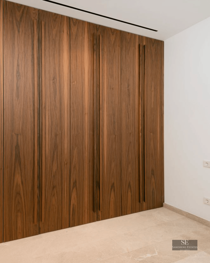 Floor-to-ceiling dark wood built-in wardrobe with vertical integrated handles in a bright room with stone floors.