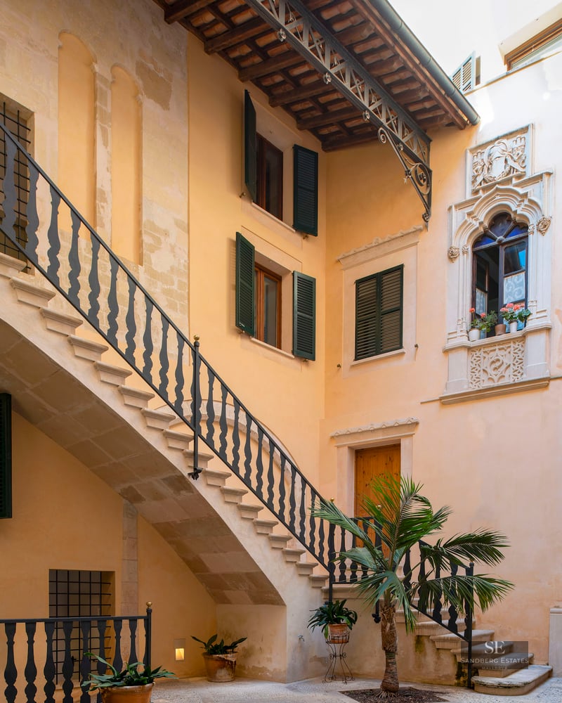 Interior courtyard featuring a stone staircase, wrought iron railings, green shutters, and a central palm tree.