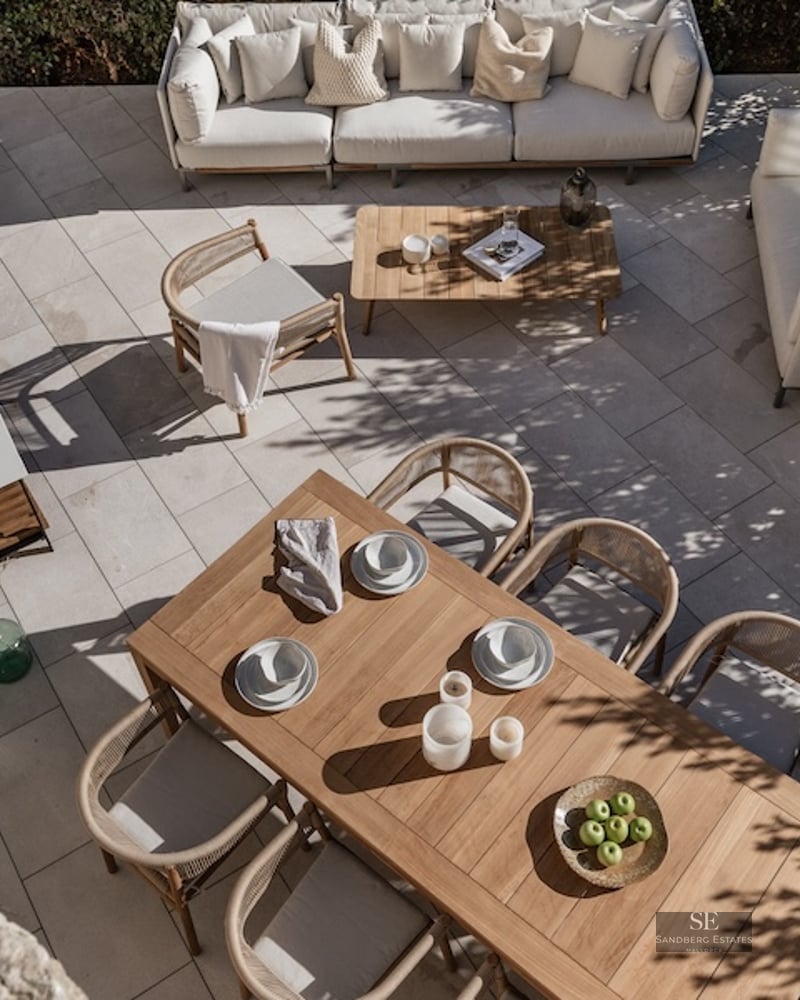 Overhead view of a modern stone patio featuring a teak dining set, white sofas, and a sleek outdoor kitchen with BBQ.