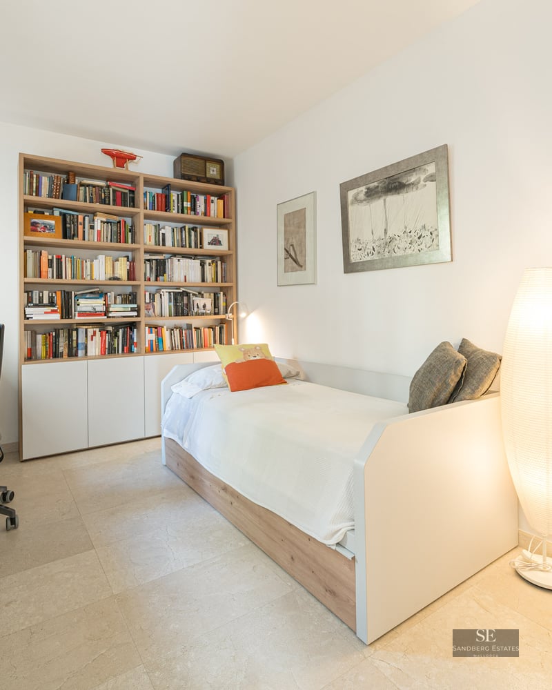Bright bedroom featuring a single bed, a large wooden bookshelf, a desk with a chair, and a glowing floor lamp.