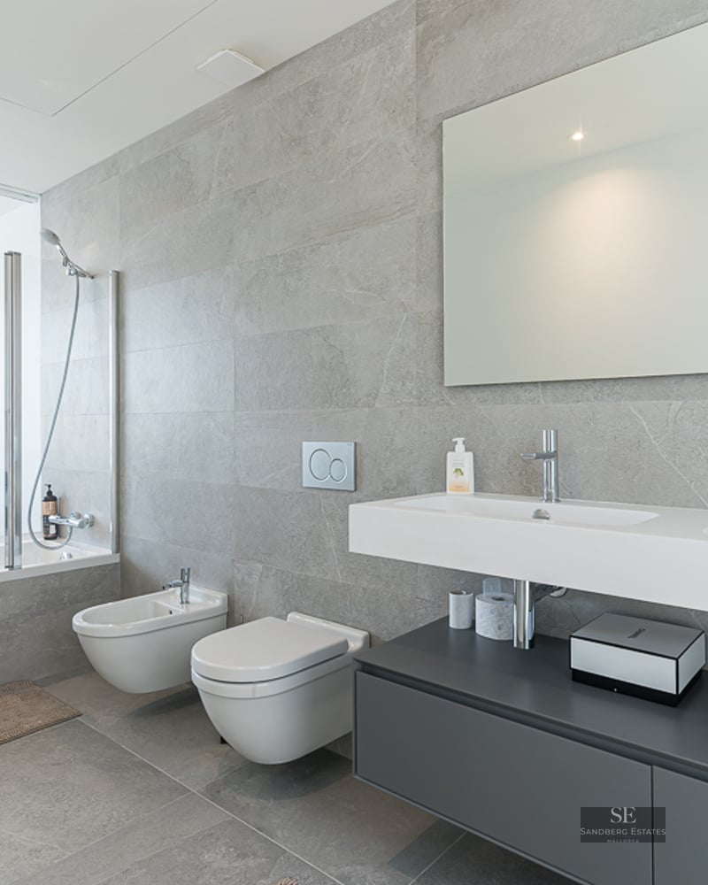 Modern bathroom featuring grey stone tiles, double vanity, wall-hung toilet, and a glass wall looking into a bedroom.