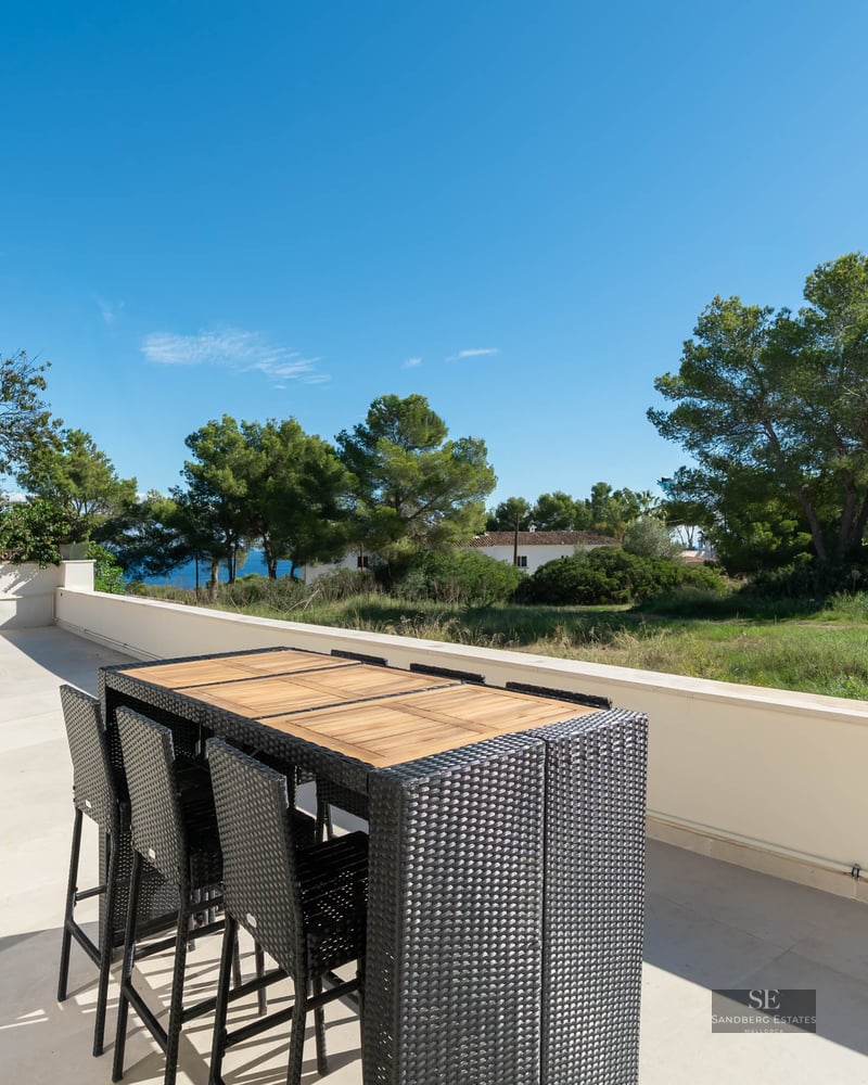 Terrasse ensoleillée avec table haute en rotin et vue sur les pins et la mer sous un ciel bleu dégagé.