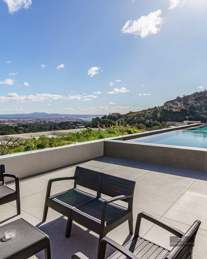 Luxury terrace featuring an infinity pool, glass railings, black outdoor furniture, and a panoramic city and sea view.