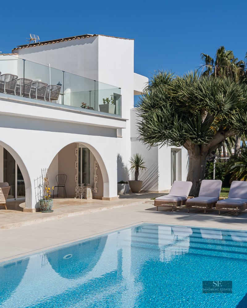 White arched villa overlooking a turquoise swimming pool with sun loungers and palm trees under a clear blue sky.