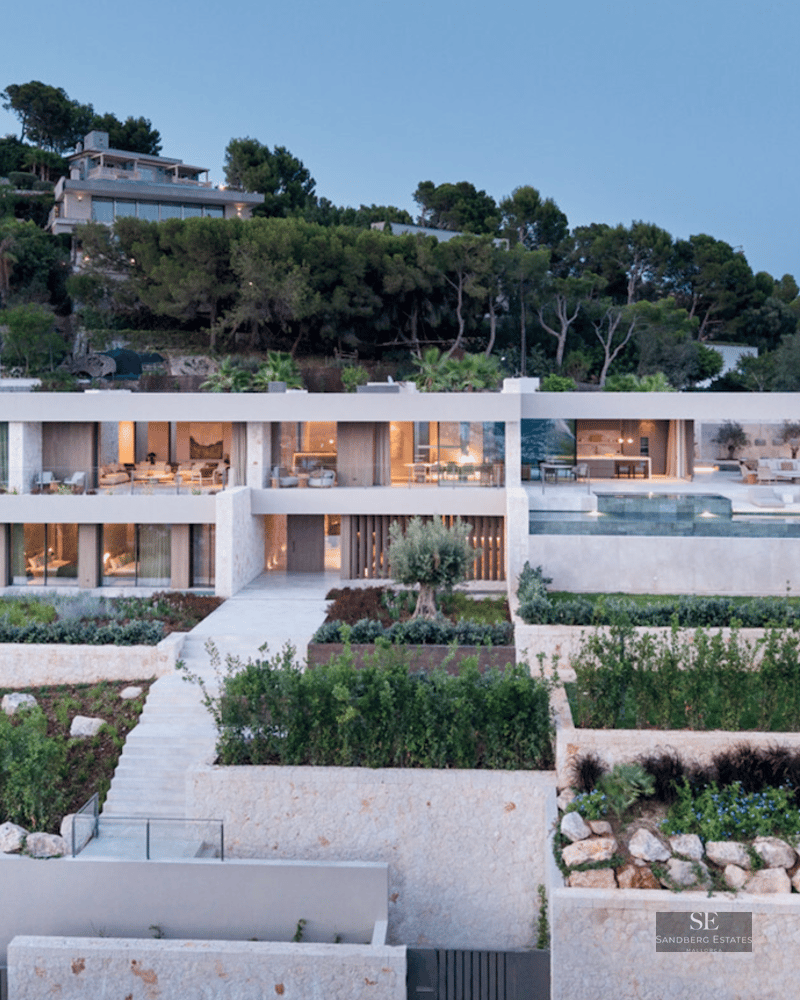 Modern two-story villa with infinity pool, stone terraced gardens, and illuminated glass facade at dusk.