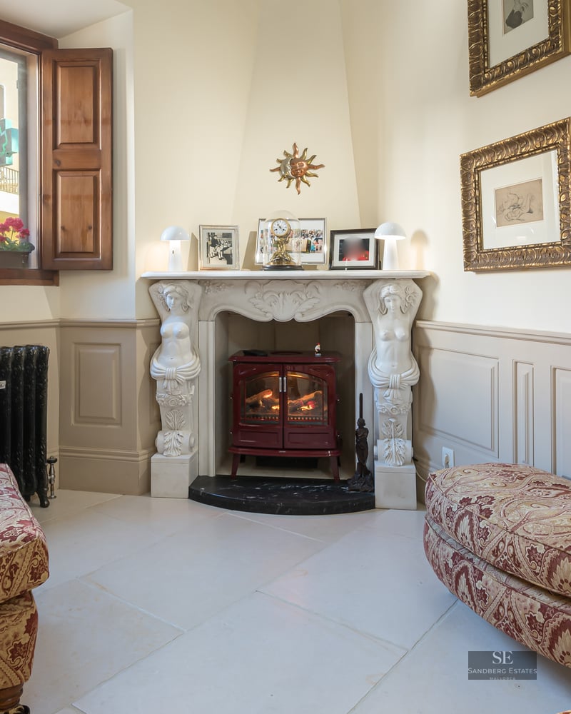 A cozy sitting area featuring an ornate white fireplace with statues, a red stove, and damask-patterned armchairs.