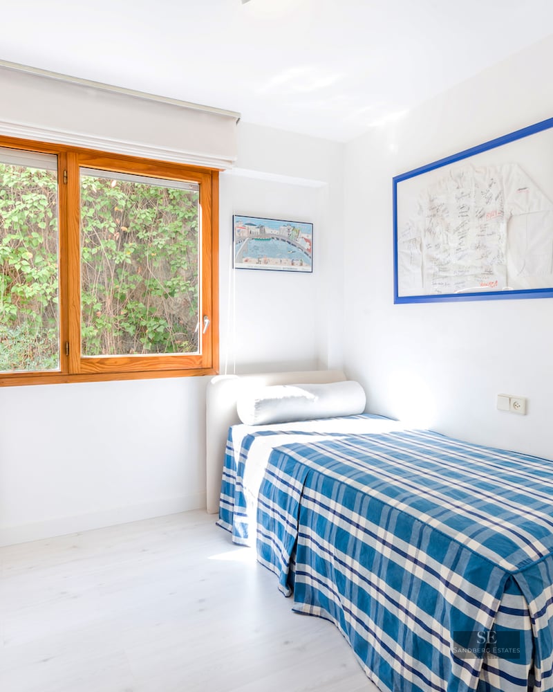 Bright single bedroom featuring a blue checkered bed, white desk, and a large wooden window looking onto green foliage.
