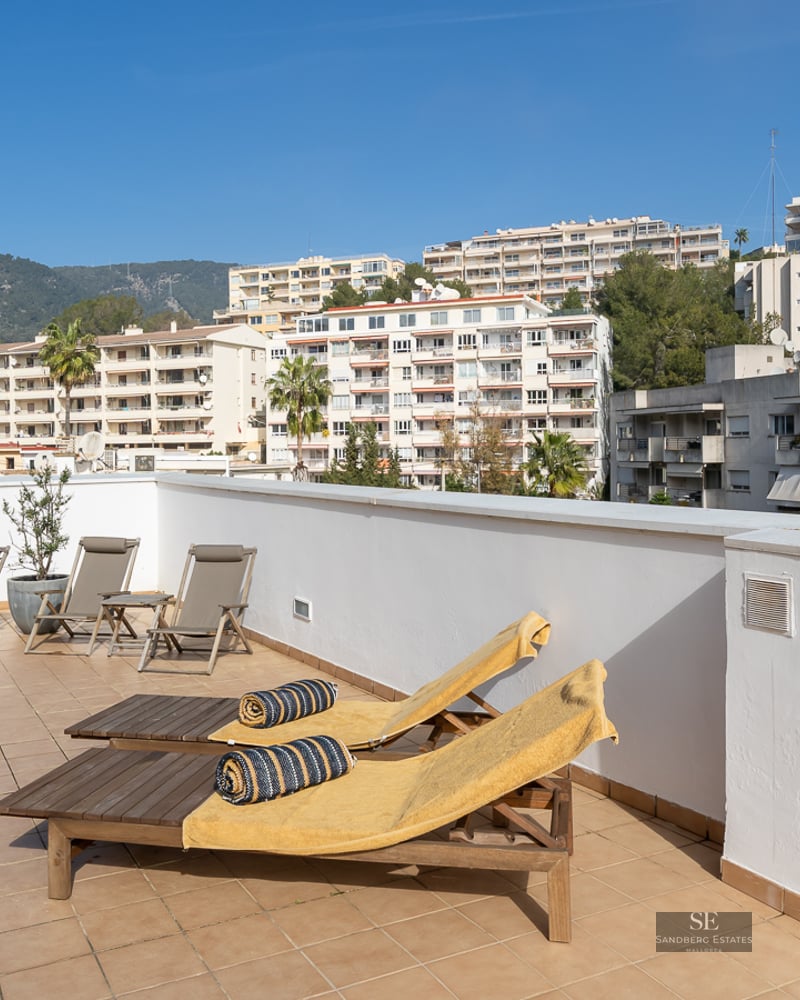 Spacious rooftop terrace featuring wooden sun loungers, tiled flooring, and views of urban buildings and mountains.