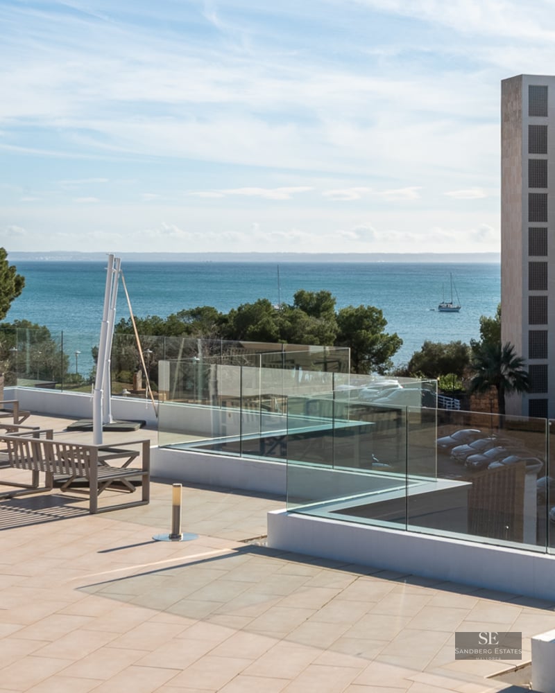 Grande terrasse carrelée avec garde-corps en verre et meubles en bois, surplombant la mer bleue et les bâtiments.