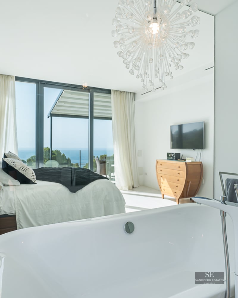 Modern master suite featuring a white freestanding bathtub in the foreground and a large bed with a sea view.
