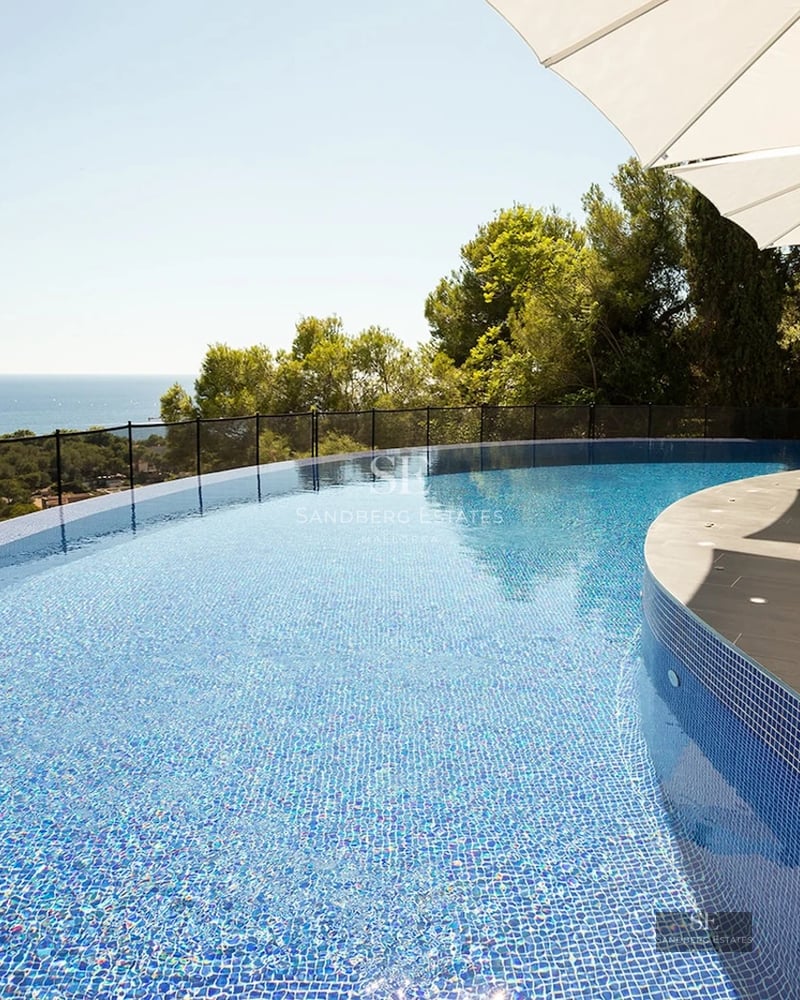 A curved infinity swimming pool with blue mosaic tiles overlooking a green valley and the Mediterranean Sea.