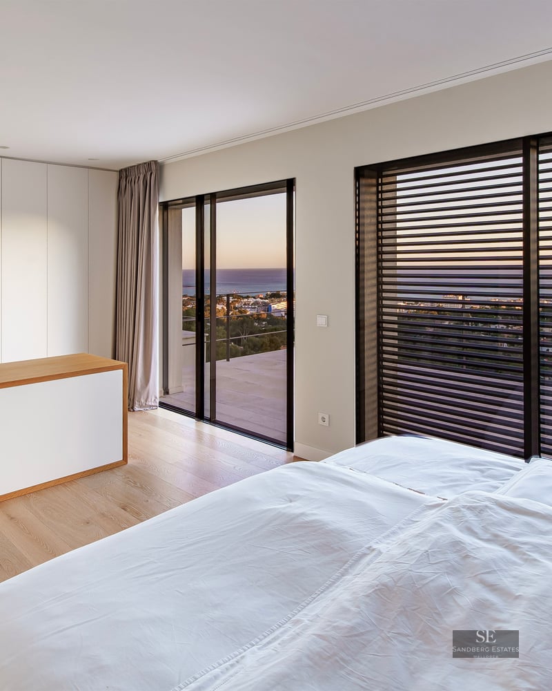 Modern bedroom with white bedding, wood floors, and large windows overlooking the sea at sunset.