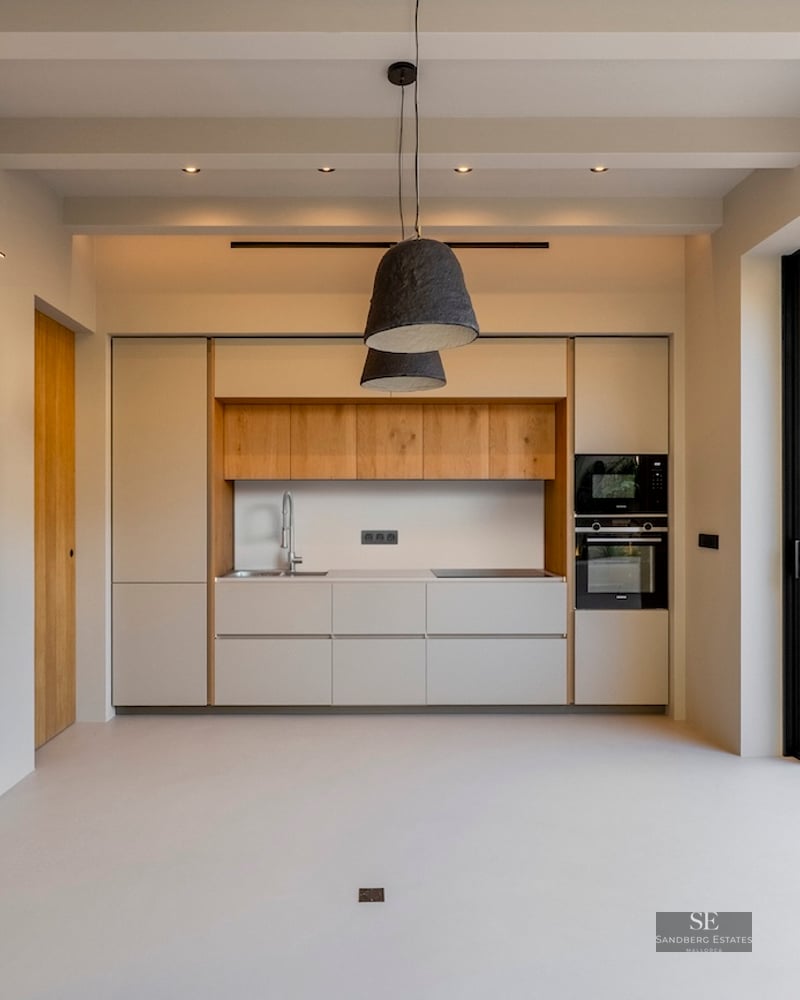 Cuisine minimaliste avec accents en bois clair, grand tableau rouge et baies vitrées ouvrant sur un patio ensoleillé.