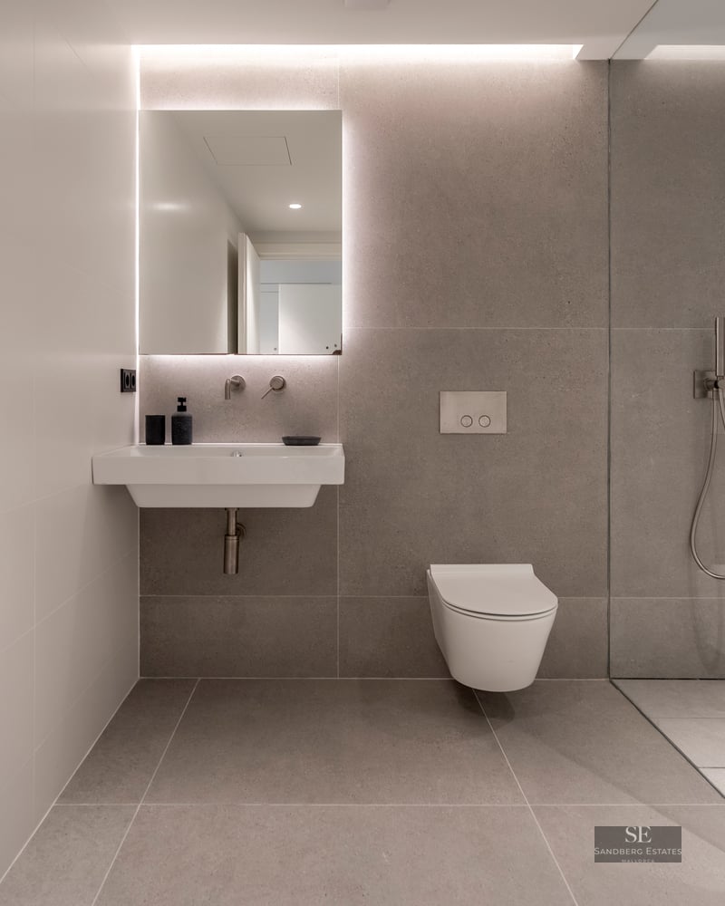 Modern bathroom featuring grey tiles, a backlit mirror above a white sink, a wall-hung toilet, and a glass shower partition.