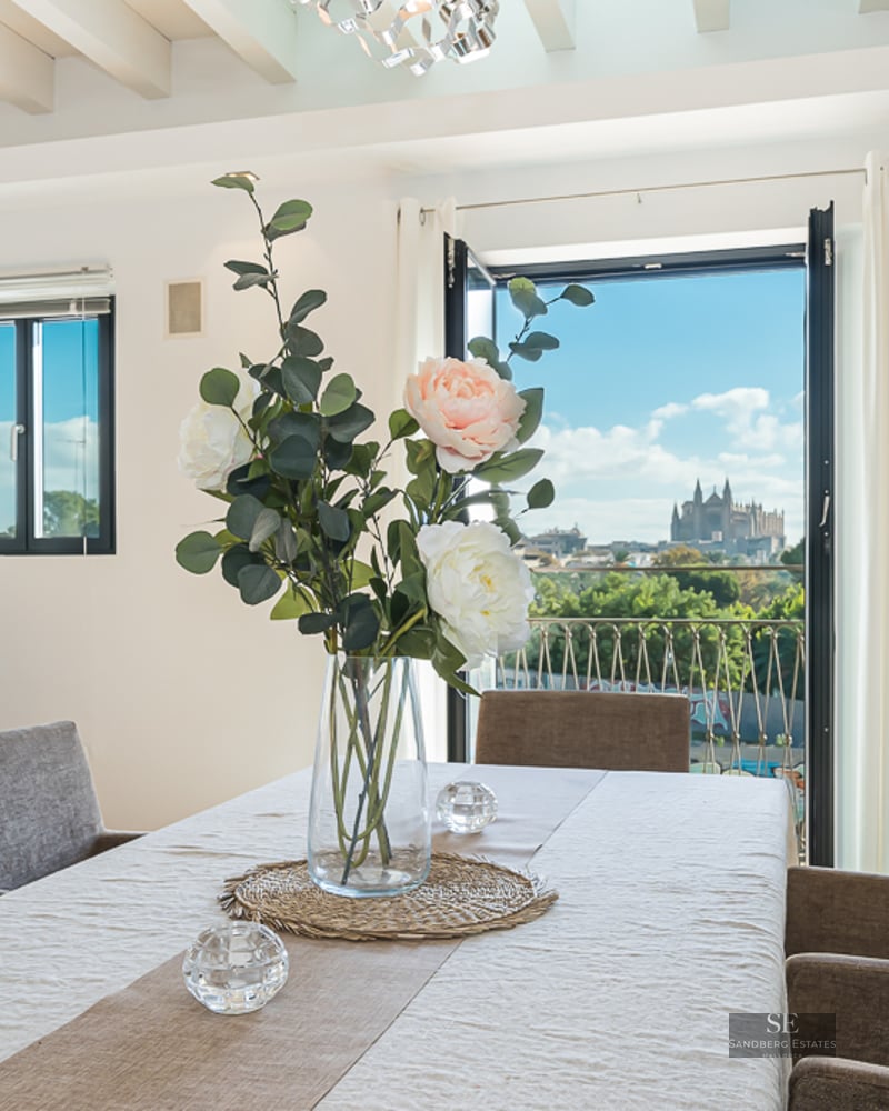 Bright dining room with white table, grey chairs, and a view of Palma Cathedral through a balcony door.