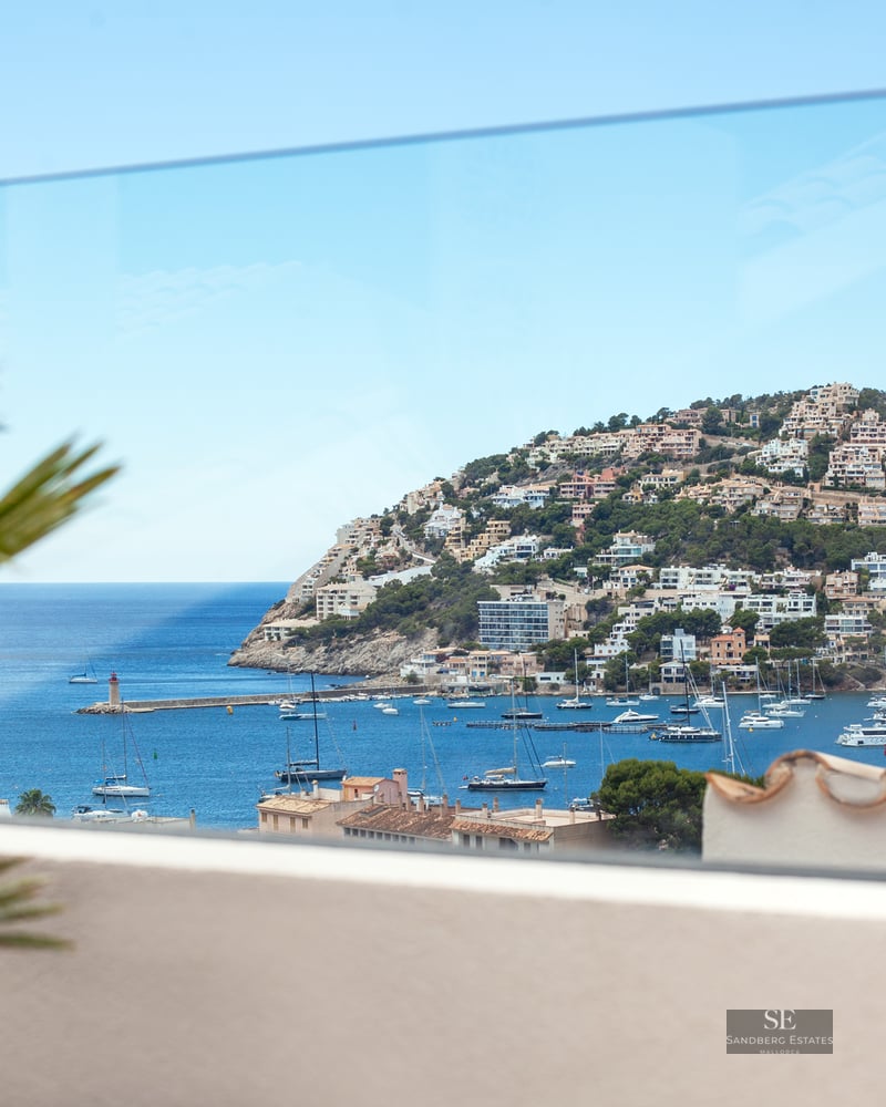 View through a glass balcony railing of a Mediterranean harbor filled with boats and a hillside town.