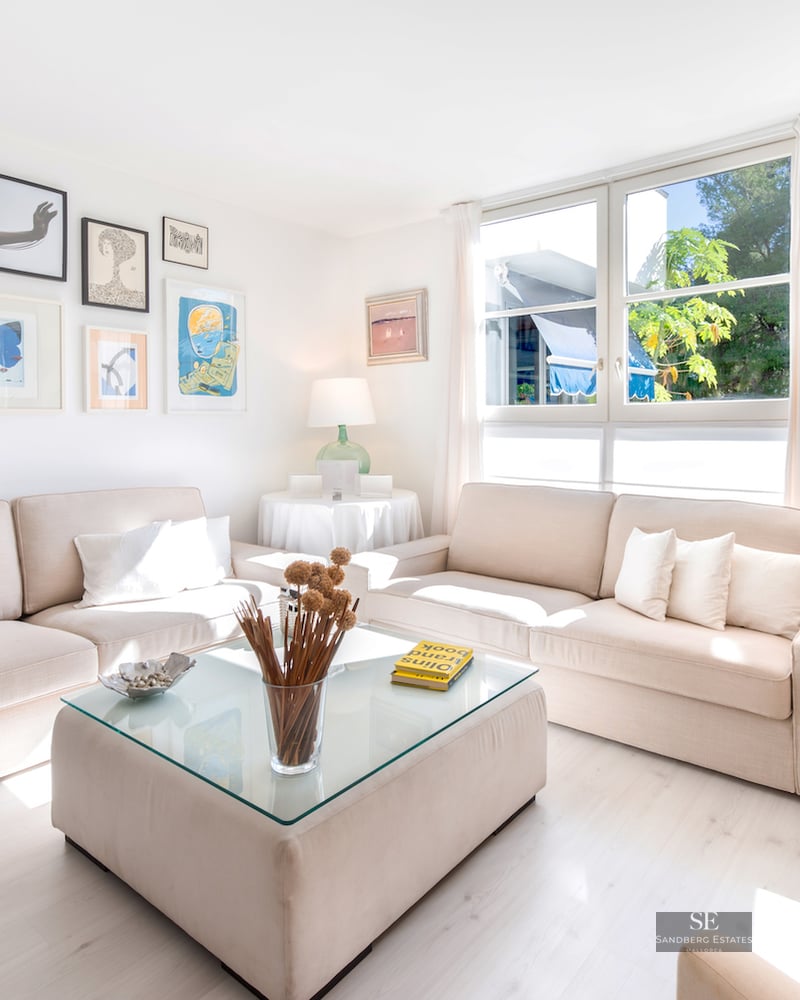 Bright living room with two beige sofas, a glass-top coffee table, large windows, and a wall of framed art.