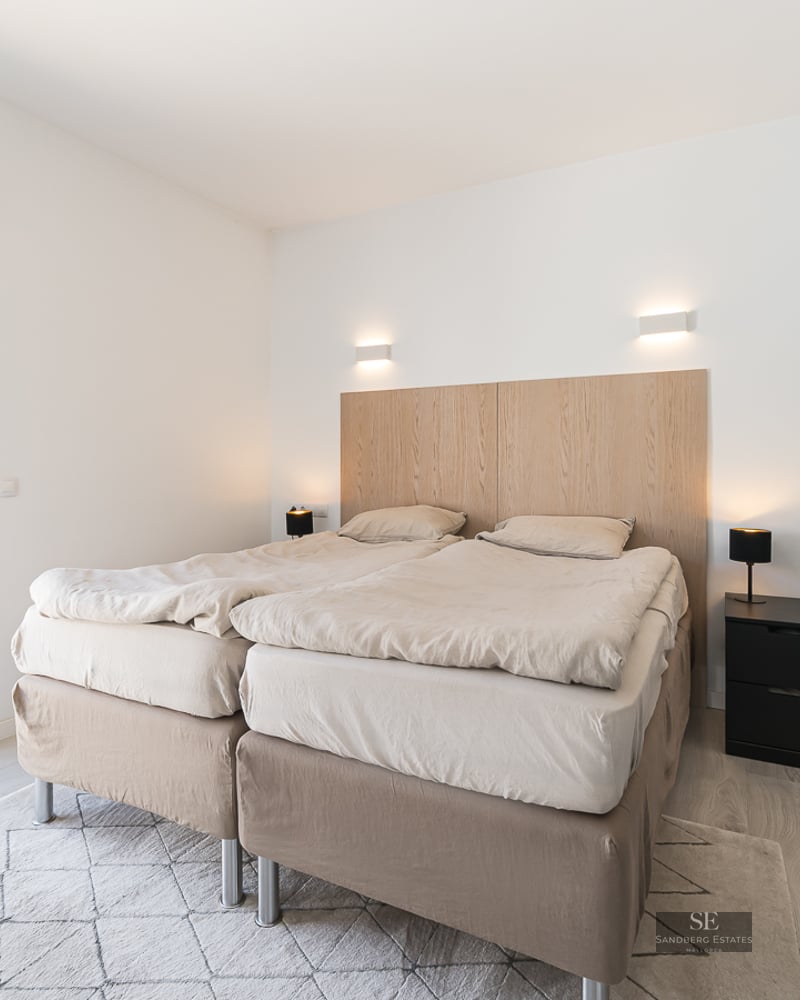 Bright bedroom featuring a double bed, light wood headboard, and a sliding glass door opening to a sunny balcony.