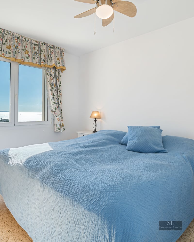 A bright bedroom with a blue bedspread, sea views through a floral-curtained window, and bedside lamps.