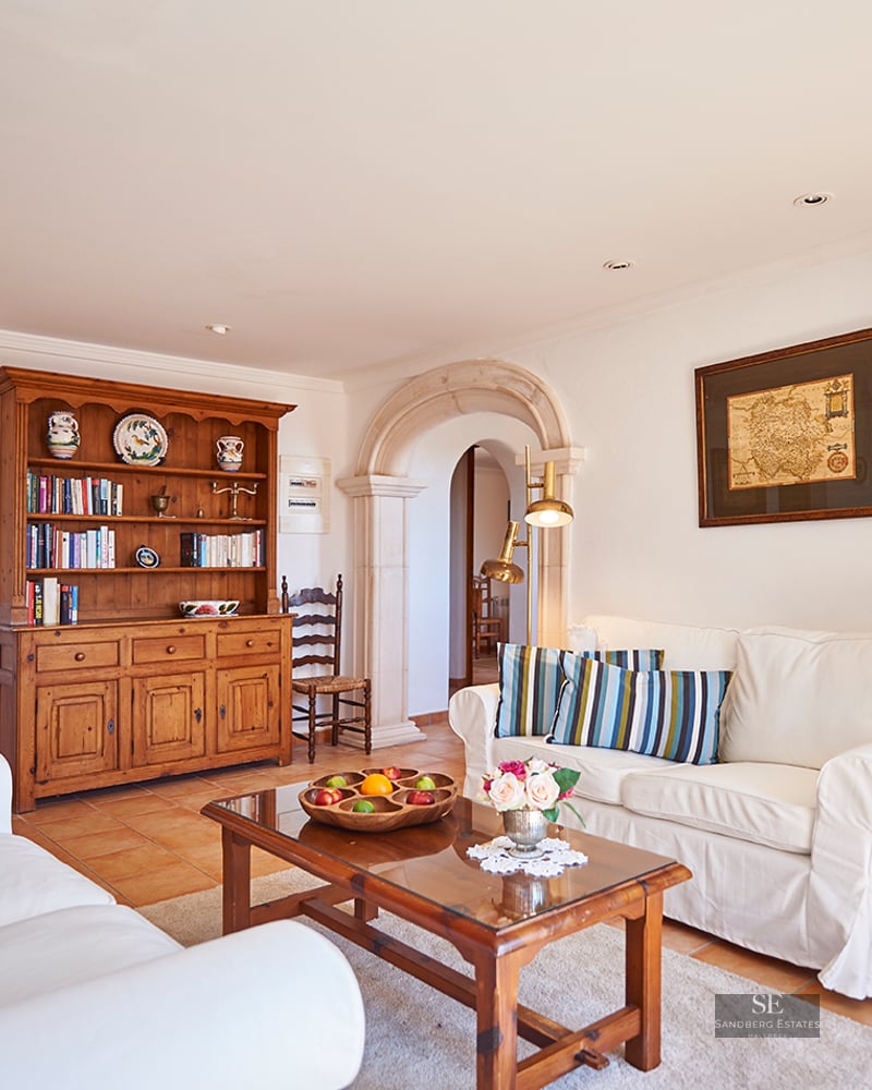Bright living room featuring white sofas, terracotta tile floors, wooden furniture, and stone arches leading to a kitchen.