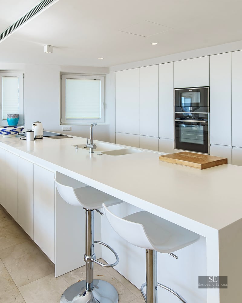 A bright, modern white kitchen featuring a large central island with bar stools and integrated appliances.