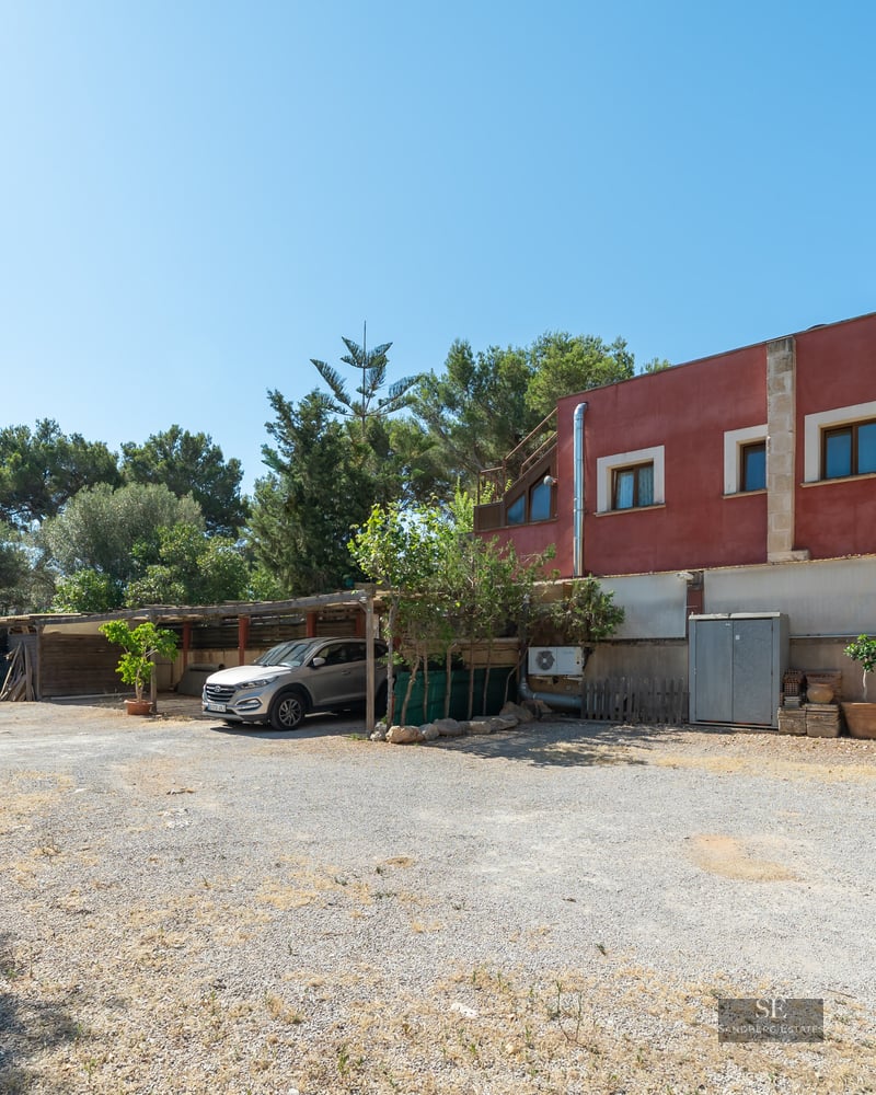 A wide gravel parking area in front of a red building with a silver car parked under a wooden carport.
