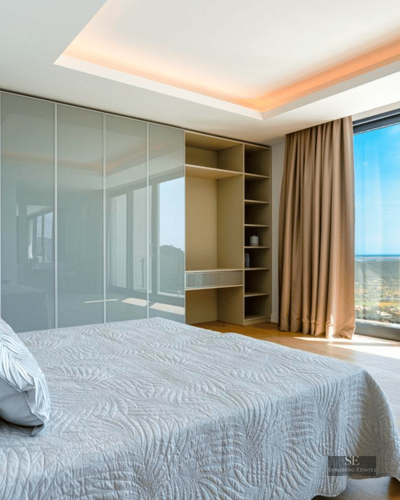 Modern bedroom with large floor-to-ceiling windows overlooking a coastal bay, featuring built-in closets and wooden floors.