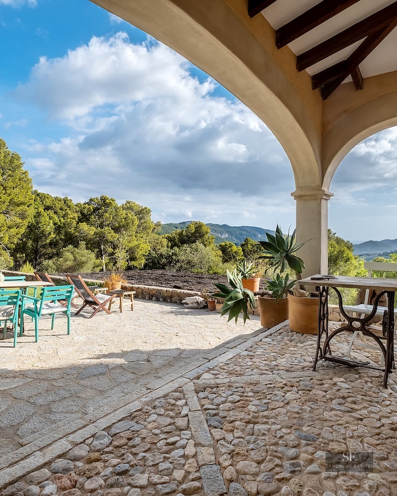 Stone terrace with arches overlooking a lush green valley and mountains, featuring teal and wood outdoor furniture.
