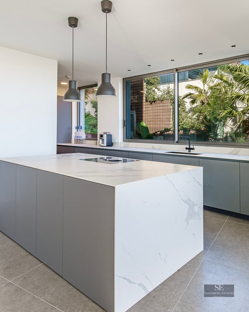 Modern kitchen with a large white marble island, grey cabinetry, and a window overlooking lush tropical plants.