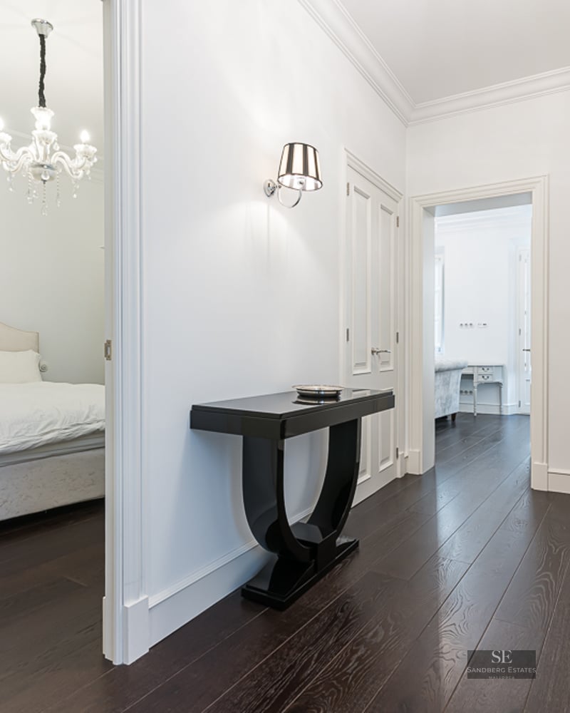 Bright hallway with dark wood floors, black console table, and view into a bedroom with a crystal chandelier.