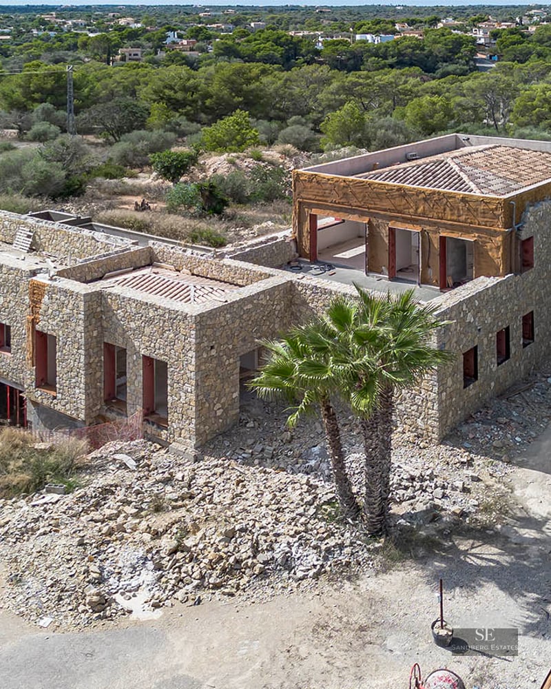 Elevated view of a large stone-walled luxury villa currently under construction surrounded by Mediterranean vegetation.
