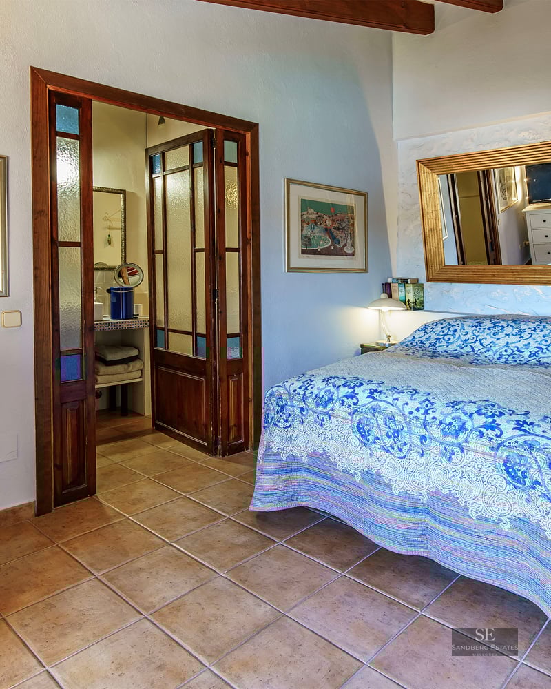 Bedroom with blue patterned bedspread, terracotta tile floor, large gold mirror, and dark wood doors to a bathroom.