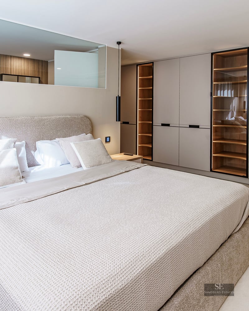 Modern master bedroom with large upholstered bed, integrated wooden wardrobes with LED lighting, and neutral decor.