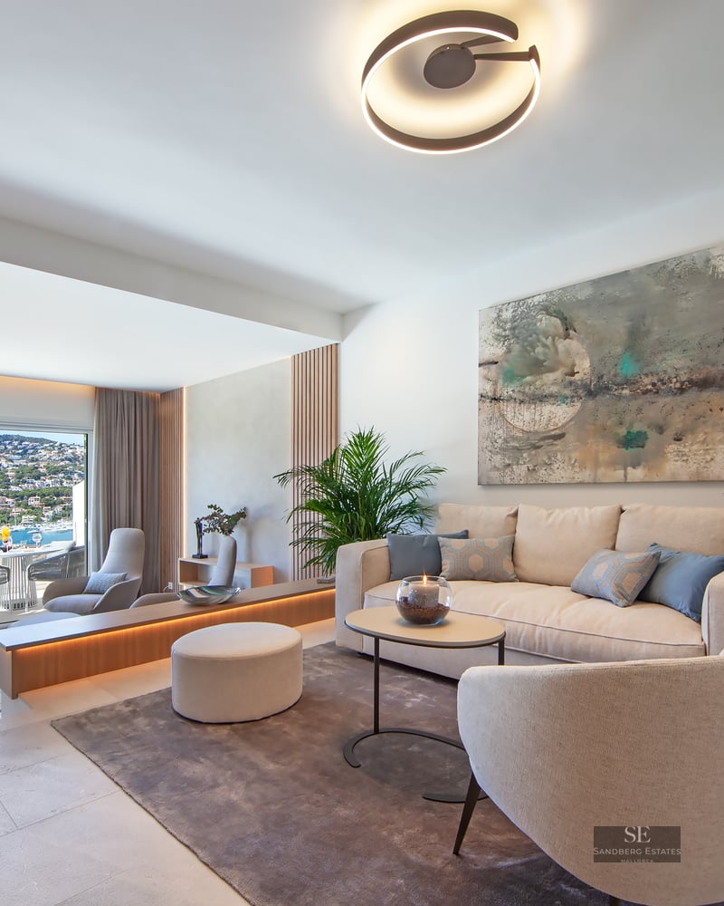 Bright modern living room with cream furniture, large windows overlooking the sea, and lush indoor plants.