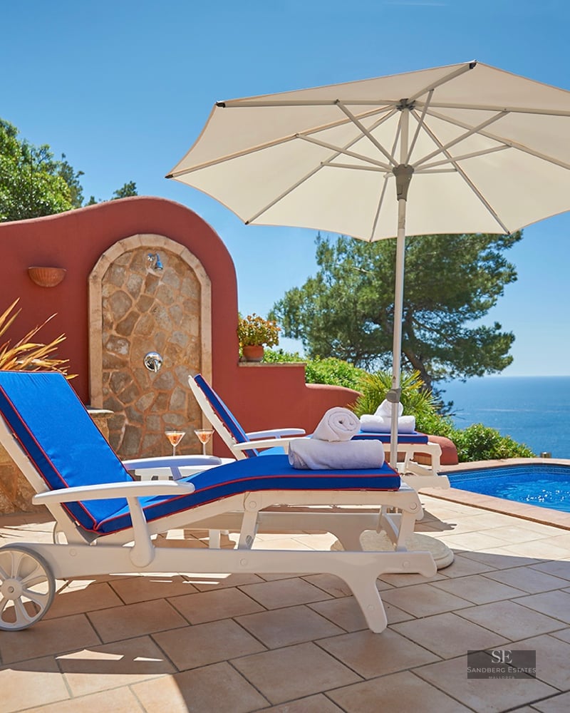 White sun loungers with blue cushions beside an infinity pool overlooking the Mediterranean Sea and cliffs.