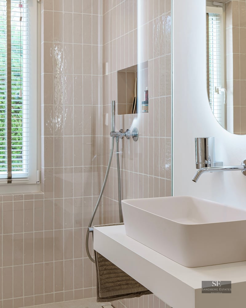 A clean, modern bathroom featuring a white vessel sink, backlit oval mirror, and soft beige vertical tiling.