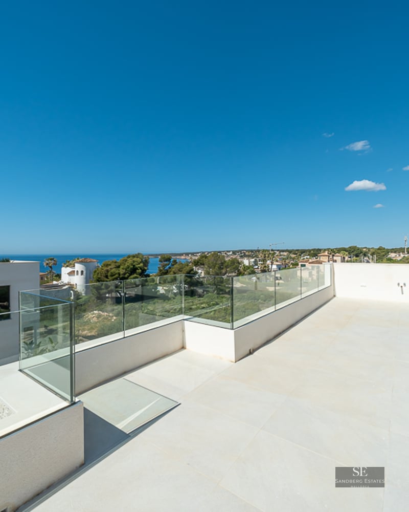 A spacious modern rooftop terrace with glass railings overlooking a coastal landscape and the blue sea under a clear sky.
