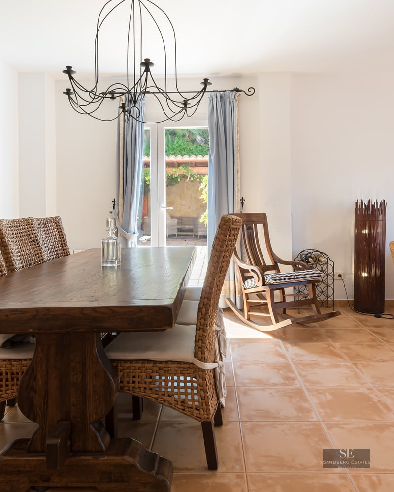 Spacious dining area with a solid wood table and wicker chairs on terracotta tiles, leading to a sunlit lounge area.