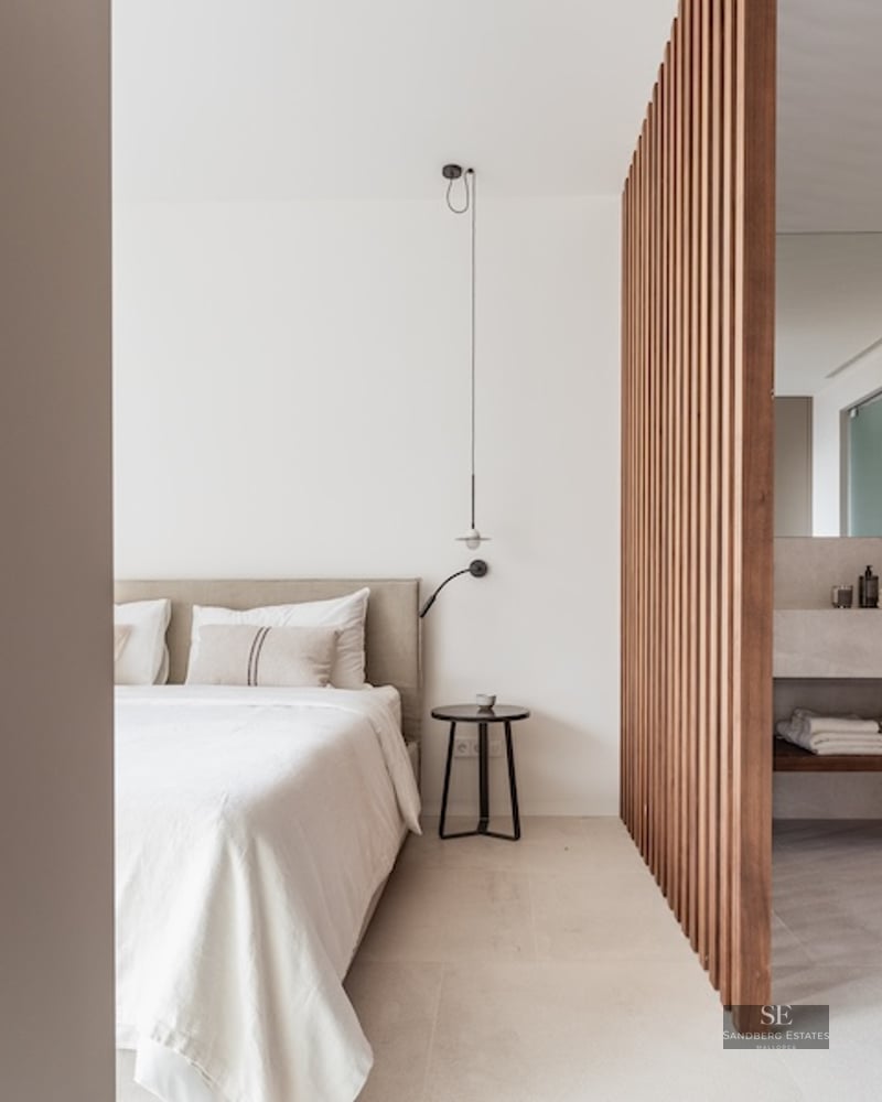 Modern bedroom with a beige bed and a wooden slat divider separating a stone vanity bathroom area.