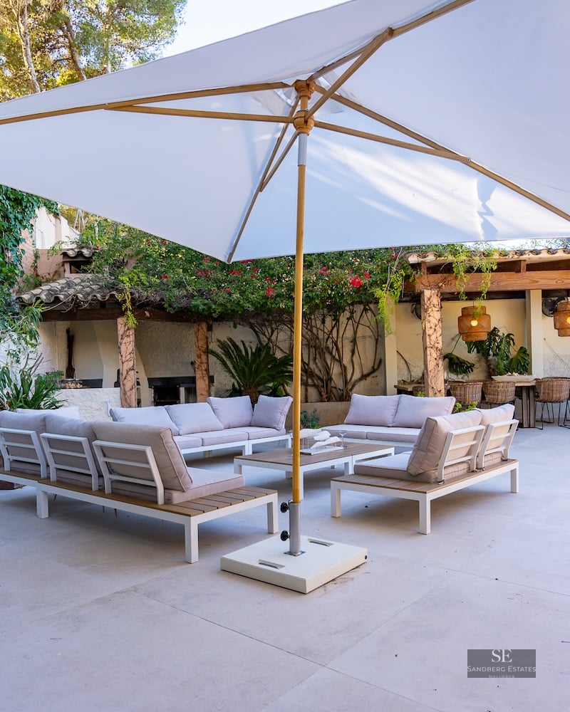 Spacious terrace featuring modern sofas, a large white parasol, and a dining area under a pergola surrounded by greenery.