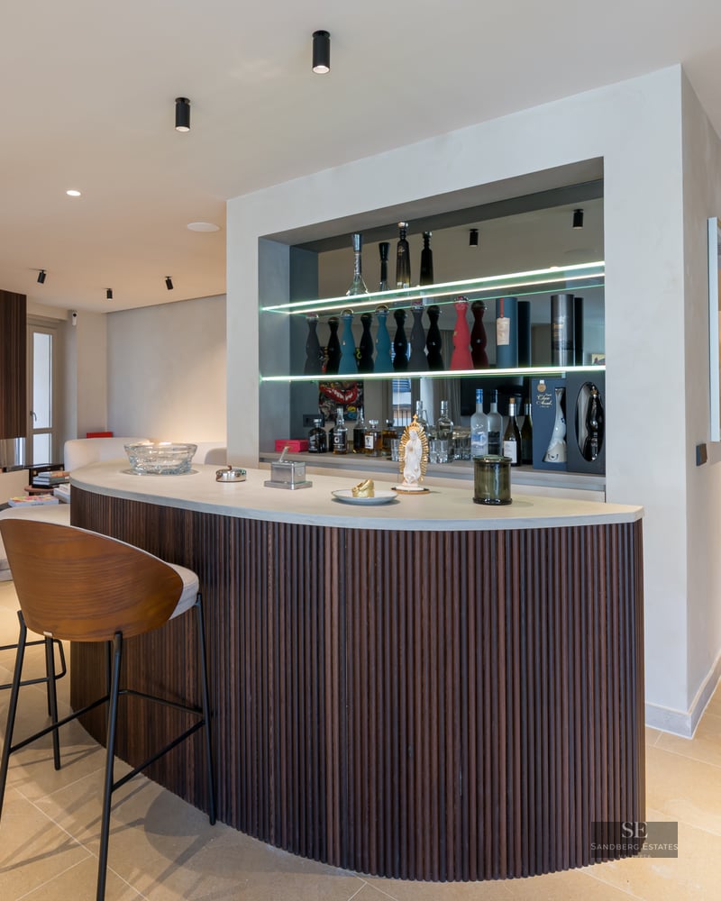 Curved home bar with dark wood slats, two designer stools, and backlit shelves with premium liquor bottles.