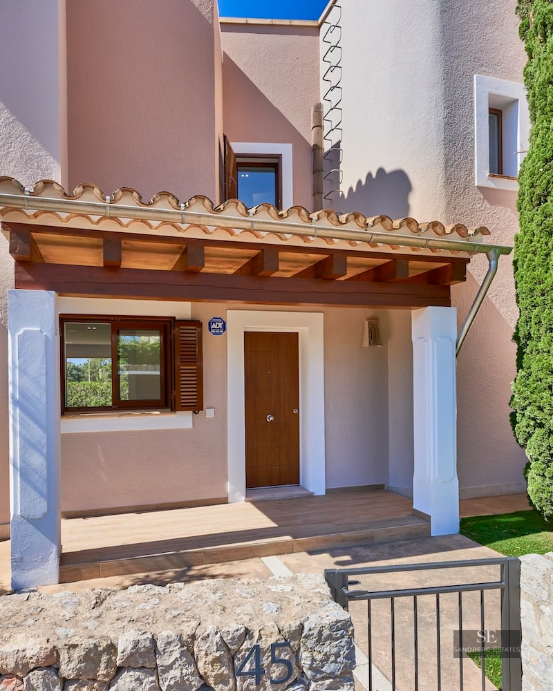 Entrance of a beige villa with a wooden door, stone wall, and a tall cypress tree under a bright blue sky.