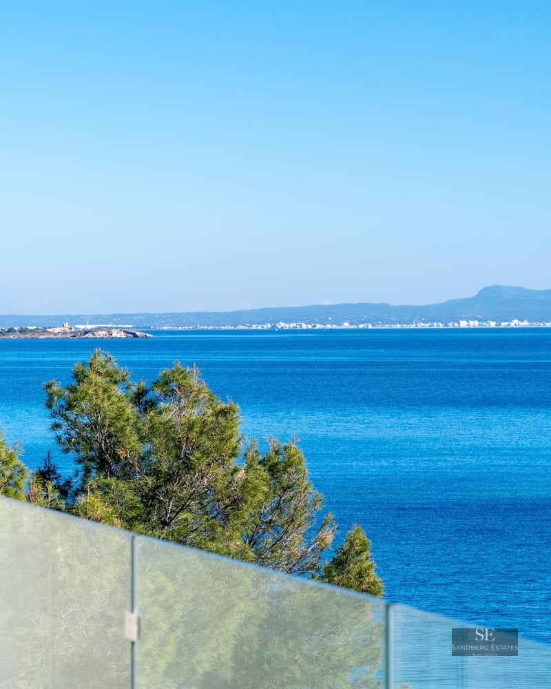 View of the deep blue Mediterranean sea and distant coastline from a terrace with a glass railing and pine trees.