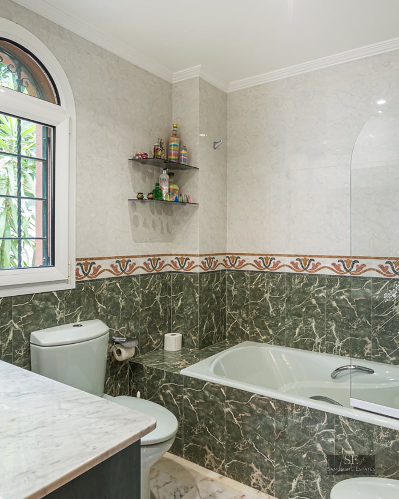 A bathroom featuring green and white marble tiling, a bathtub with a glass screen, and a bright arched window.