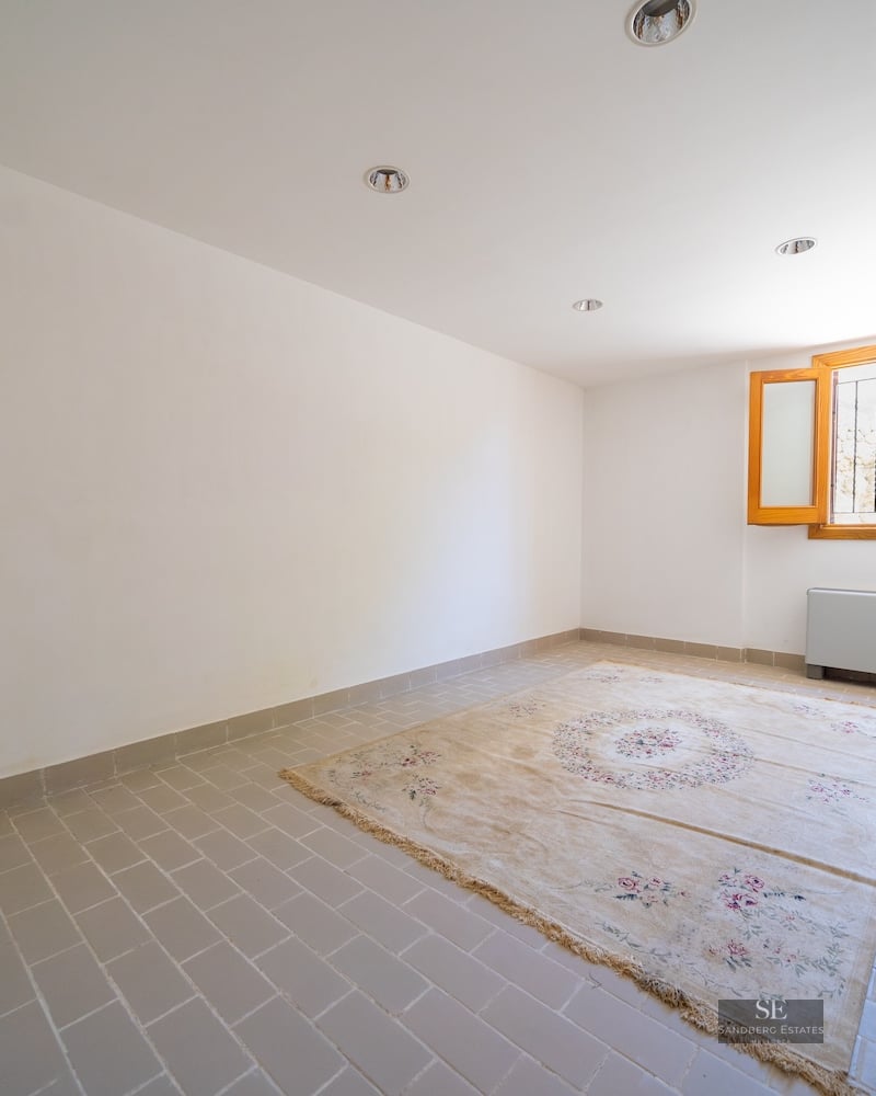 An empty room with light grey floor tiles, white walls, a patterned rug, and a small wooden window.