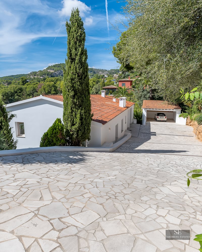 White stone paved driveway leading to a Mediterranean style villa with terracotta roof and mountains in background.