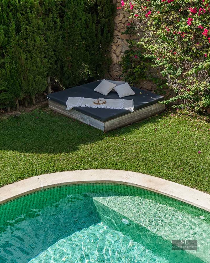A green-tiled swimming pool next to a manicured lawn and a daybed with pillows under pink bougainvillea flowers.