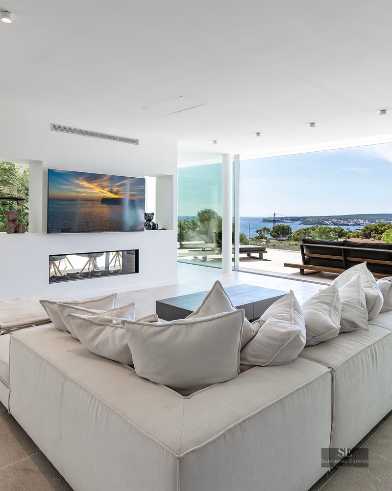 Spacious modern living room with large white sectional sofa, integrated fireplace, and floor-to-ceiling windows with sea views.